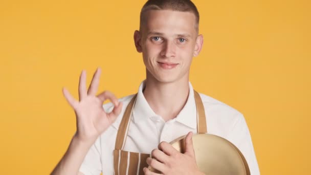 Young handsome waiter in uniform with tray happily showing ok and bellissimo gesture on camera over colorful background - Footage, Video