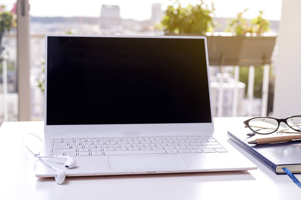 Laptop na biurku ze słuchawkami, notatnikiem i okularami. Biuro z pięknym widokiem na słoneczny dzień. Równowaga życia zawodowego - Zdjęcie, obraz