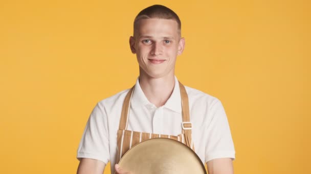 Joven camarero atractivo en uniforme con bandeja sonriendo alegremente en la cámara sobre fondo colorido - Metraje, vídeo