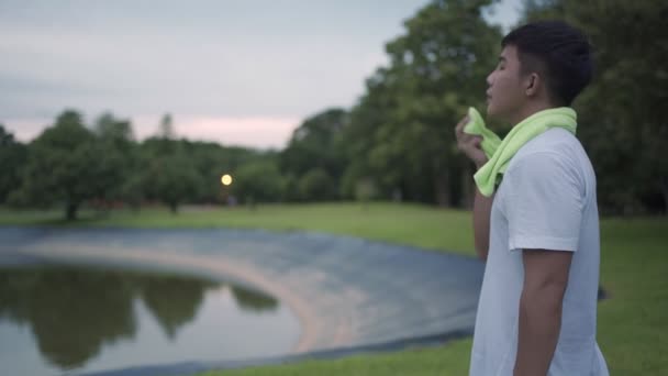 Asiatischer Mann trinkt Wasser nach dem Sport im Park mit Bäumen und Teich im Hintergrund, steht nach dem Sport entspannt im Freien, Happy active man Lifestyle, aufgenommen bei Sonnenuntergang am Ende des Tages - Filmmaterial, Video