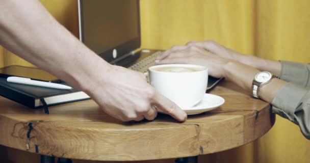 Close-up van de handen van een mannelijke ober serveert een mok hete cappuccino aan een vrouwelijke klant die werkt op een laptop in een cafe. - Video