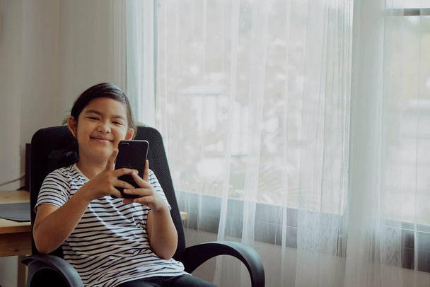 Asiática niña usando smartphone para videollamada en una silla en casa
 - Foto, imagen