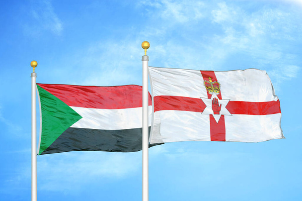 Σουδάν και Βόρεια Ιρλανδία δύο σημαίες σε κοντάρια σημαίας και φόντο μπλε συννεφιασμένο ουρανό - Φωτογραφία, εικόνα