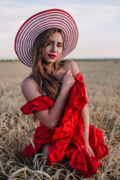 Beautiful girl in red dress and striped hat posing in a wheat field - Fotó, kép