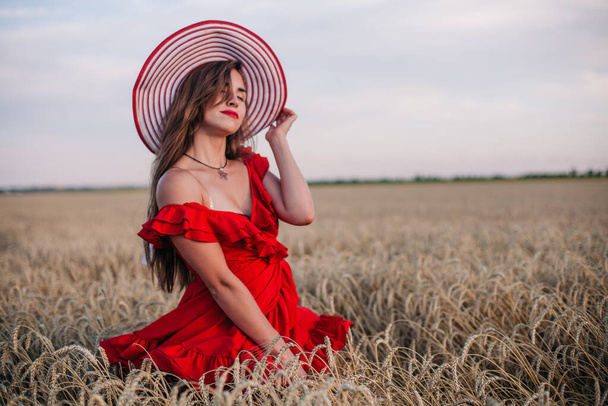 Красивая девушка в красном платье и в полосатой шляпе позирует на пшеничном поле - Фото, изображение