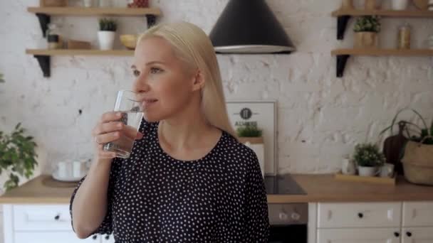 The girl drinks water from a glass standing in the bright kitchen. - Záběry, video