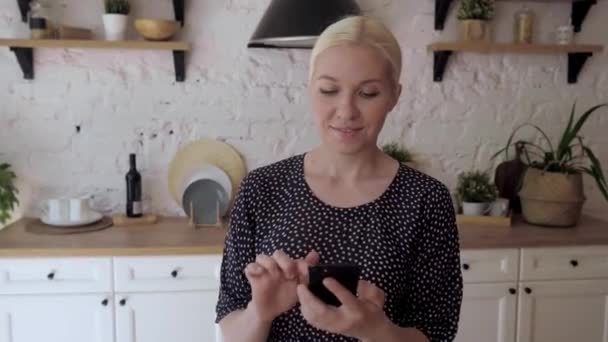 Žena stojící v kuchyni drží chytrý telefon vychutnat vzdálený chat s přáteli - Záběry, video