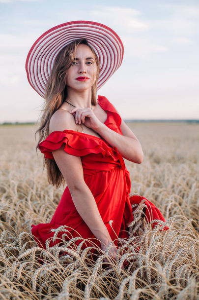 Beautiful girl in red dress and striped hat posing in a wheat field - Photo, image