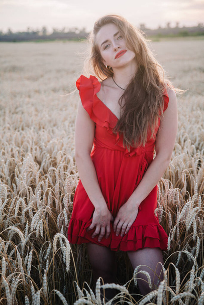 Beautiful girl in red dress and striped hat posing in a wheat field - Valokuva, kuva
