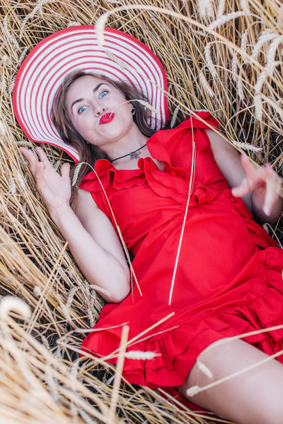 Beautiful girl in red dress and striped hat posing in a wheat field - Фото, изображение