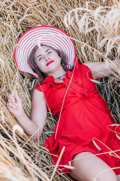 Beautiful girl in red dress and striped hat posing in a wheat field - Фото, зображення