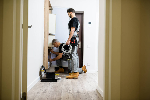 Full length shot of aged electrician, repairman in uniform working, installing ethernet cable or router in fuse box while his young colleague watching him, holding cable - Zdjęcie, obraz