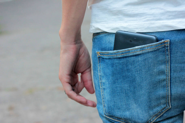 Młoda kobieta w niebieskich dżinsach ze smartfonem w tylnej kieszeni. Zbliżenie czarnej komórki w kieszeni. - Zdjęcie, obraz