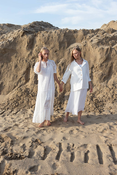 twee aantrekkelijke jonge tweeling zusters poseren bij zandgroeve in elegante witte kleren - Foto, afbeelding