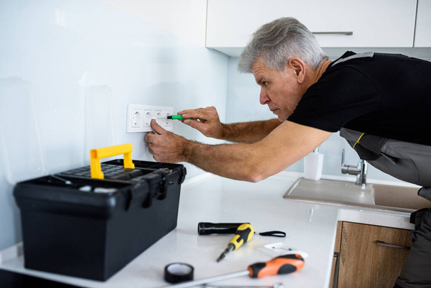 The best quality service. Aged electrician, repairman in uniform working, installing or fixing socket in the kitchen using screwdriver. Repair service concept - Foto, afbeelding