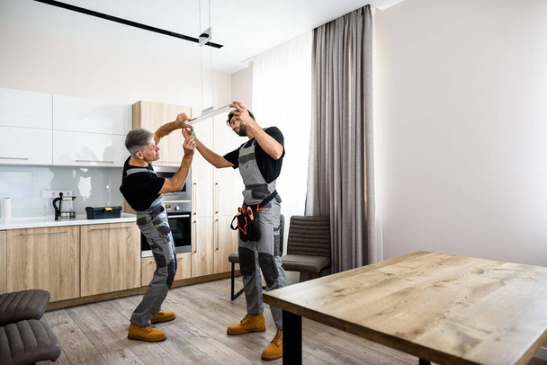 Dlouhý záběr dvou elektrikářů, pracovníků v uniformách instalujících elektrickou lampu, světlo uvnitř bytu v kuchyni. Koncept dekorace stavby - Fotografie, Obrázek