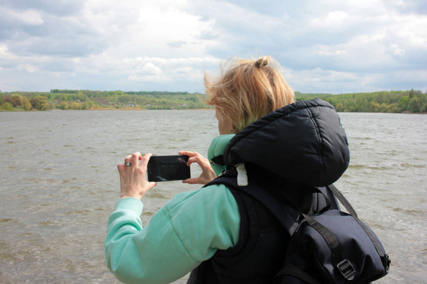 Jeune femme blonde prenant une photo du paysage estival, printanier ou automnal avec rivière et arbres à l'horizon. Vue arrière - Photo, image