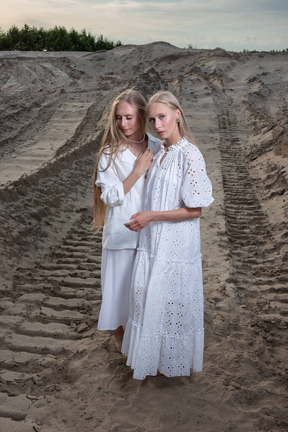 дві привабливі молоді сестри-близнюки позують на піщаному кар'єрі в елегантному білому одязі
 - Фото, зображення