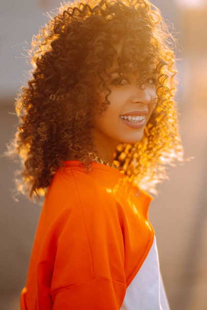 Portrait of stylish  African  American  girl - dancer at sunset. Beautiful and young woman with curly hair posing on a city street. Fashion lifestyle. - Fotografie, Obrázek