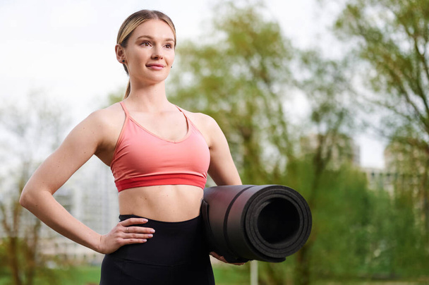 Donna fitness che indossa abbigliamento sportivo che tiene tappetino yoga all'aperto nel parco estivo - Foto, immagini