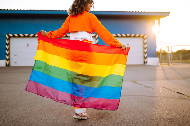 Menina lésbica afro-americana bonita com bandeira do arco-íris LGBT ao pôr-do-sol. jovem cabelo encaracolado mulher posando com lgbt orgulho bandeira
. - Foto, Imagem