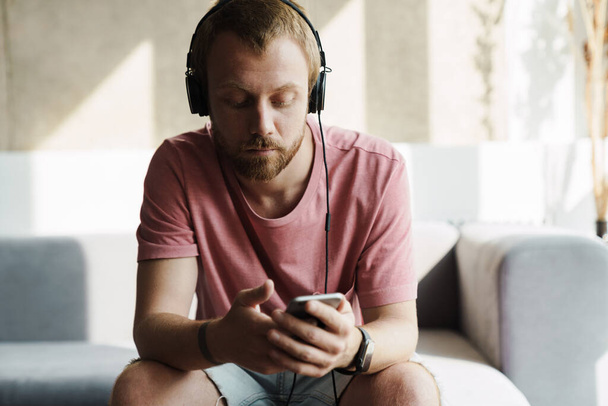 Photo of young redhead man with beard using mobile phone and headphones while sitting on sofa at home - Zdjęcie, obraz