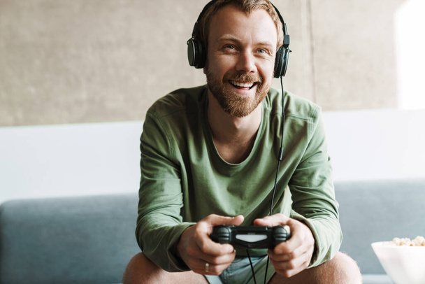 Photo of laughing man wearing headphones playing video game with joystick while sitting on sofa at home - Фото, изображение