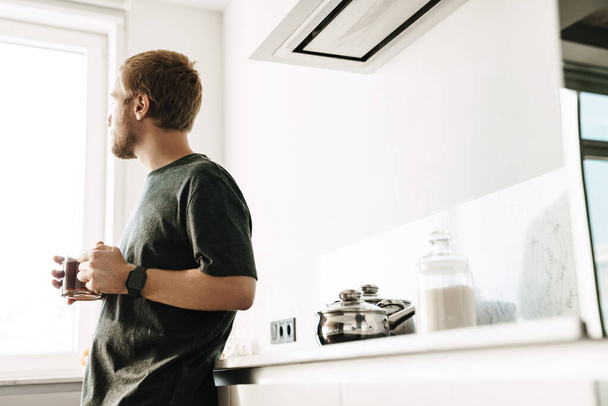 Photo of caucasian redhead man with beard drinking tea while standing in kitchen at home - Foto, Bild