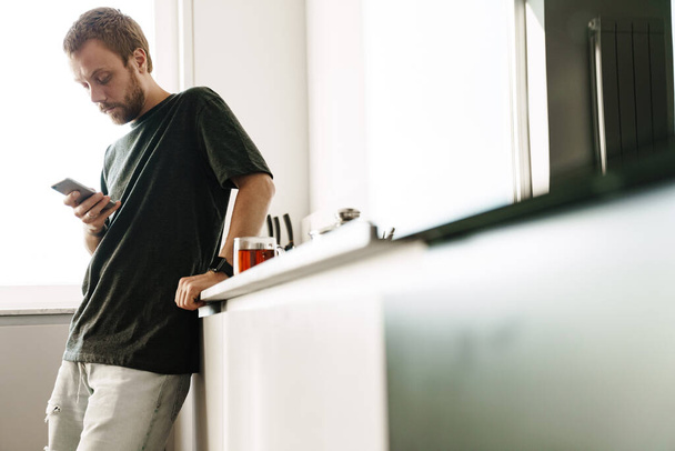 Photo of caucasian redhead man with beard using mobile phone while standing in kitchen at home - Fotó, kép