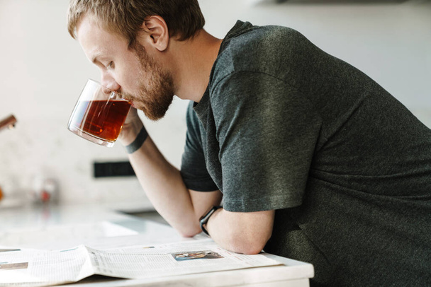 Photo of serious redhead man with beard reading newspaper while drinking tea in kitchen at home - Photo, Image