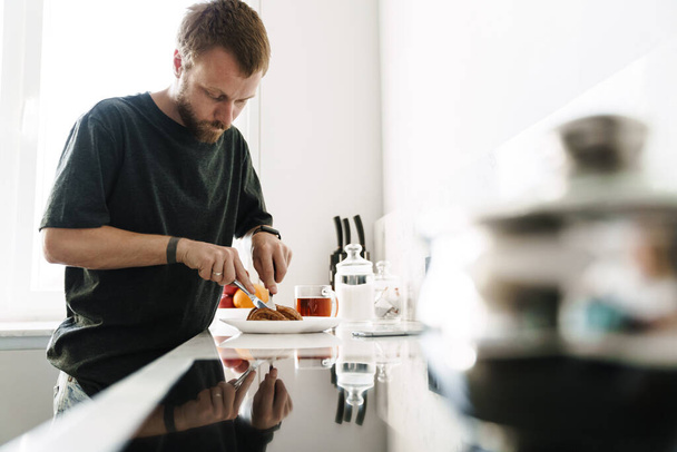 Photo of caucasian young man with beard eating croissant while having breakfast in kitchen at home - Fotografie, Obrázek