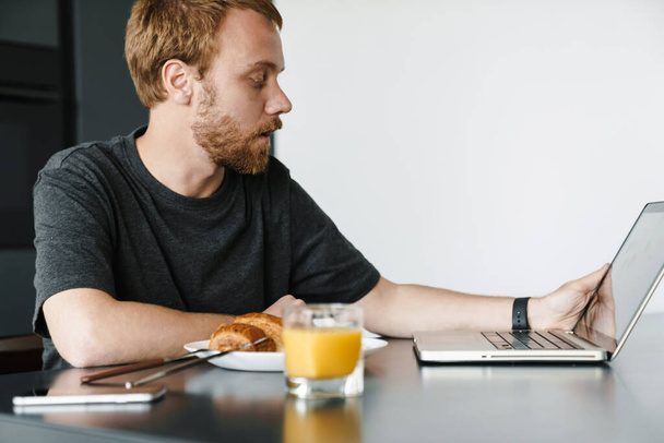 Photo of caucasian young man with beard using laptop while having breakfast at table in kitchen - Foto, Bild