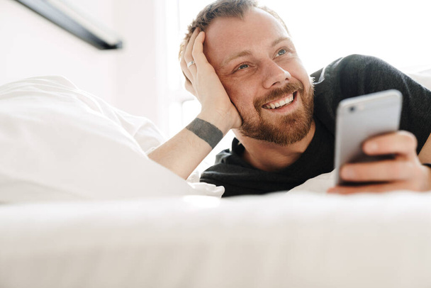 Photo of smiling bearded man using mobile phone while lying after sleep in white bed at bright room - Φωτογραφία, εικόνα
