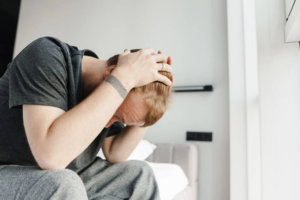 Photo of displeased redhead man grabbing his head while sitting on bed after sleep at bright room - Φωτογραφία, εικόνα