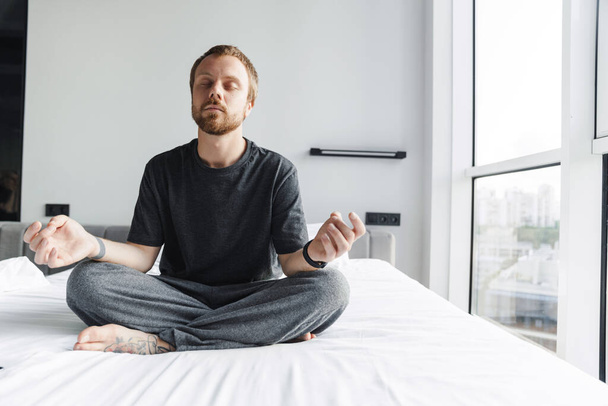 Photo of pleased redhead man meditating with eyes closed while sitting on bed at bright room - 写真・画像