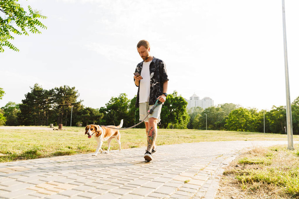 Image of young caucasian stylish man using cellphone while walking with beagle dog in summer park - Photo, image
