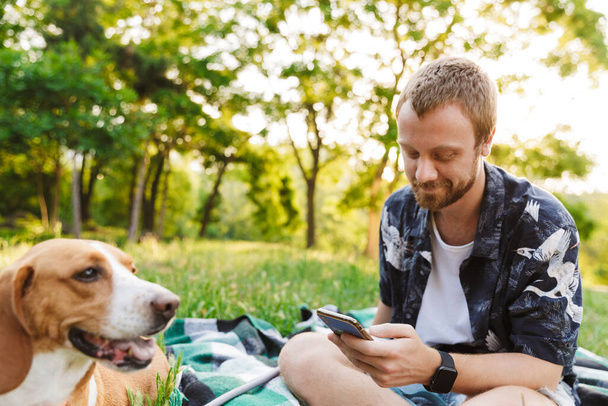 Image of young caucasian bearded man using cellphone while sitting with his beagle dog in summer park - Fotografie, Obrázek