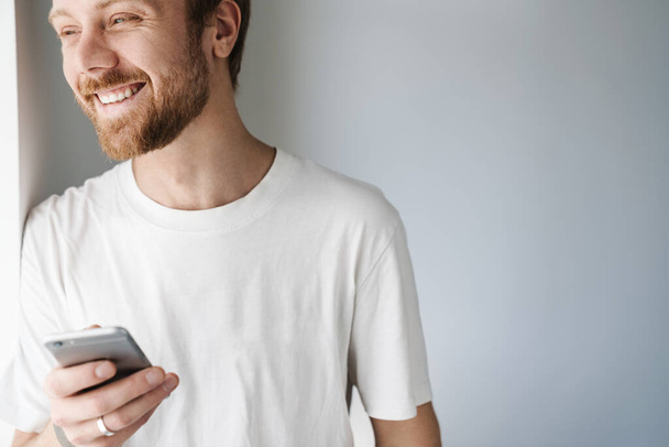 Photo of pleased bearded man with red hair using mobile phone and smiling while leaning on wall indoors - Photo, image