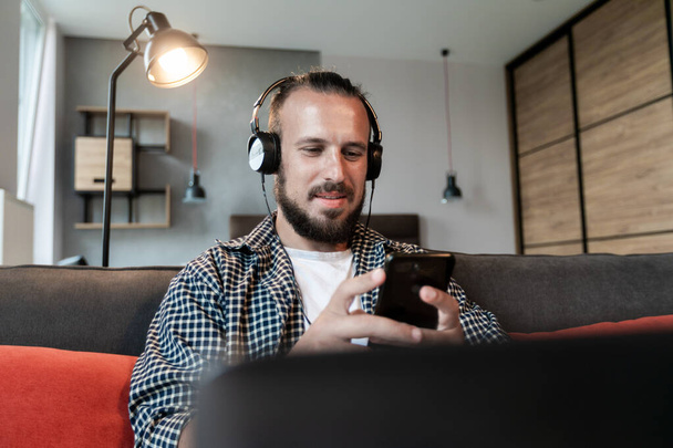 O cara ouve música em fones de ouvido, senta-se no sofá e escreve uma mensagem em seu smartphone
 - Foto, Imagem