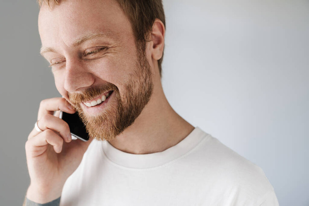 Photo of pleased bearded man with red hair talking on mobile phone and smiling indoors - Foto, afbeelding