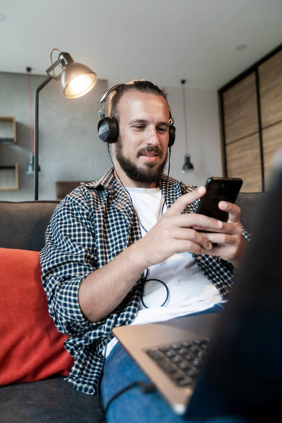 Хлопець слухає музику на навушниках, сидить на дивані і пише повідомлення на своєму смартфоні
 - Фото, зображення
