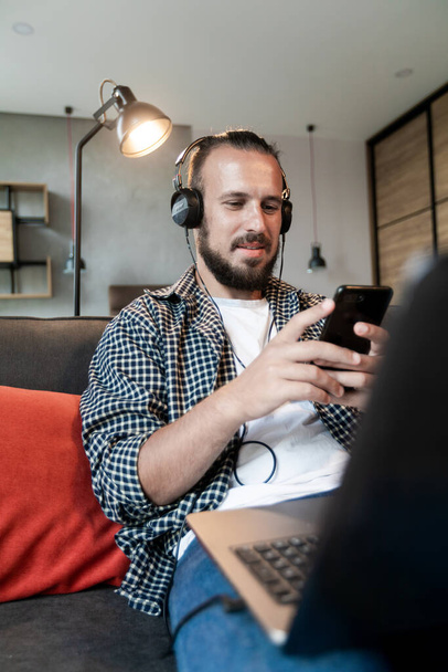 A fickó zenét hallgat fejhallgatón, leül a kanapéra és üzenetet ír az okostelefonjára. - Fotó, kép