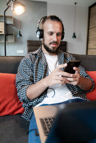 De man luistert naar muziek op de hoofdtelefoon, zit op de bank en schrijft een bericht op zijn smartphone - Foto, afbeelding
