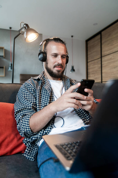 El tipo escucha música en los auriculares, se sienta en el sofá y escribe un mensaje en su teléfono inteligente
 - Foto, Imagen