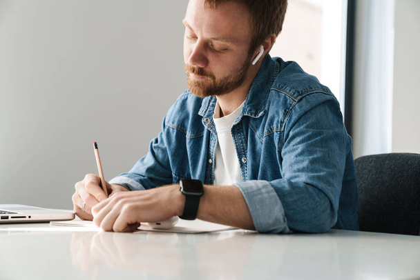 Photo of concentrated ginger man using wireless earphones while writing down notes at table indoors - Foto, Imagen