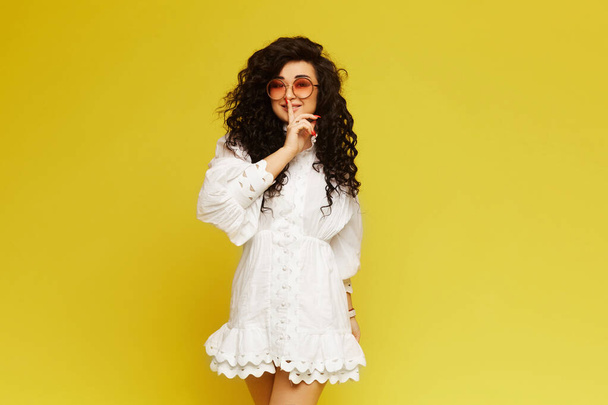 Young woman with curly hair, in a white dress and sunglasses, making gesture hush and posing on yellow background, isolated - Zdjęcie, obraz