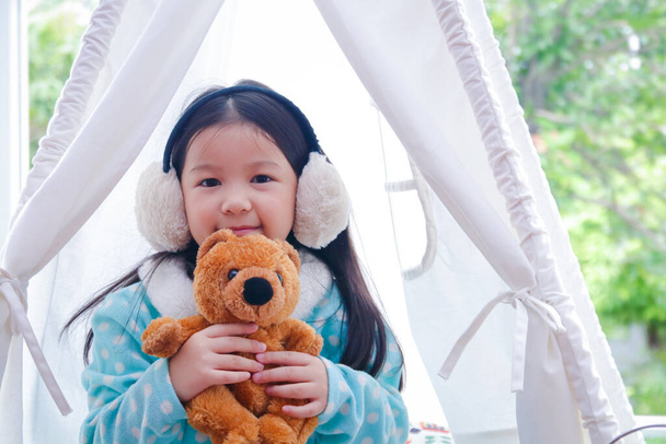 Una graziosa bambina che indossa un paraorecchie Seduta ad abbracciare un orsacchiotto marrone nella sua tenda bianca Felice nell'inverno dell'anno - Foto, immagini