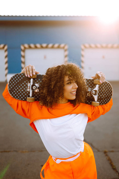 Beautiful African  American woman posing with a skateboard. Hipster girl with curly hair with skateboard at sunset. Fashion lifestyle. - Fotografie, Obrázek