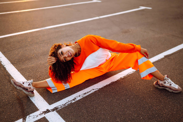Stylish girl - dancer dancing in the street at sunset. Beautiful African  American woman with curly hair in an orange suit.  Sport, dancing and urban culture concept. - Фото, изображение