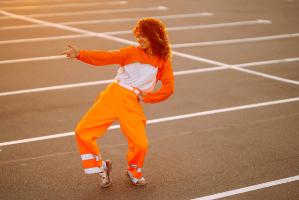 Stylish girl - dancer dancing in the street at sunset. Beautiful African  American woman with curly hair in an orange suit.  Sport, dancing and urban culture concept. - Foto, Imagen